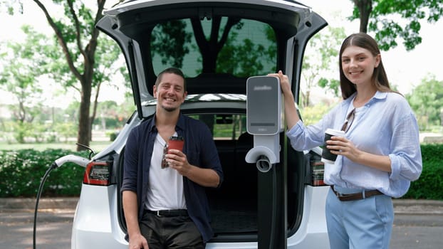 EV car recharging electricity for battery by lovey couple traveling during their road trip travel by eco friendly electric vehicle in national park and greenery forest on holiday. Exalt