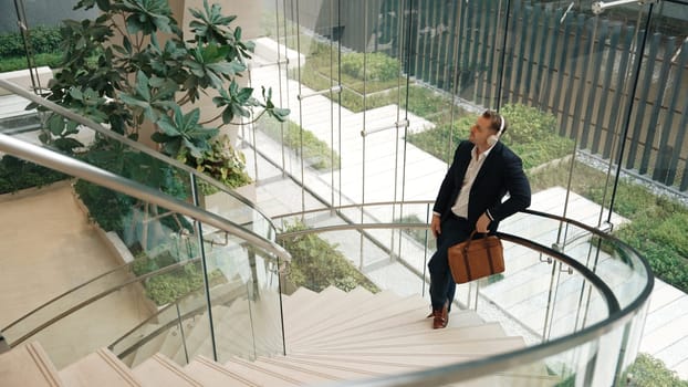 Top view of caucasian businessman listening relaxing music wearing headphone while standing at spiral stair cases at modern building. Project manager enjoy listening song while move to rhyme. Urbane.