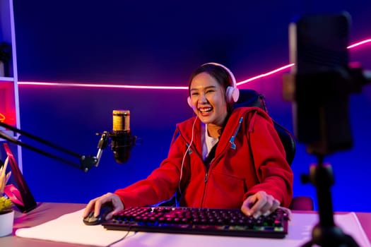 Host channel of smiling beautiful Asian girl streamer playing online game wearing headphone talking with viewers media online recording phone. Esport skilled team players in neon blue room. Stratagem.