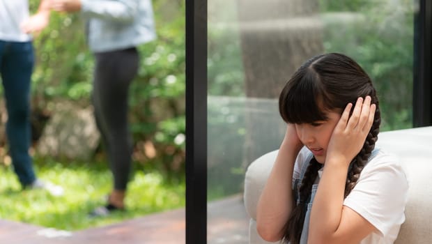 Stressed and unhappy young girl huddle in corner, cover her ears blocking sound of her parent arguing in background. Domestic violence at home and traumatic childhood develop to depression. Synchronos