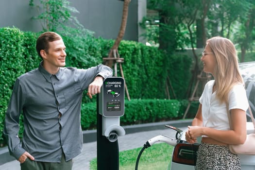 Young couple travel with EV electric car charging in green sustainable city outdoor garden in summer shows urban sustainability lifestyle by green clean rechargeable energy of electric vehicle innards