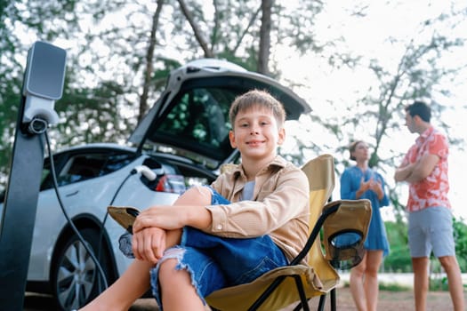 Little boy portrait sitting on camping chair with his family in background. Road trip travel with alternative energy charging station for eco-friendly car concept. Perpetual