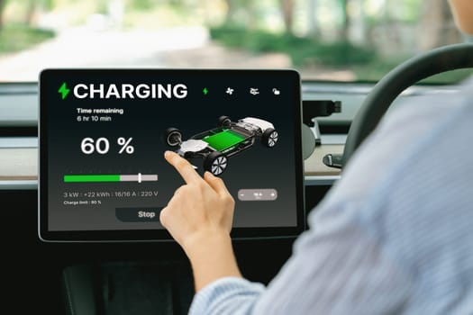 Young woman inside the vehicle, checking EV vehicle's application on battery recharging electricity status display on smart monitor screen in modern EV car on her road trip journey. Exalt