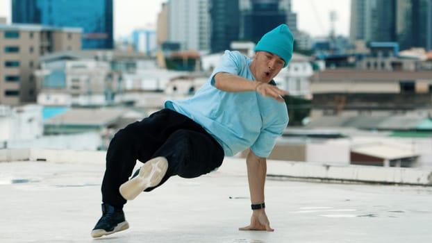 Skilled caucasian B-boy dancer practicing street dancing at rooftop with city sites or urban. Motion shot of young man performing street dance by doing freeze pose. Outdoor sport 2024. Endeavor.