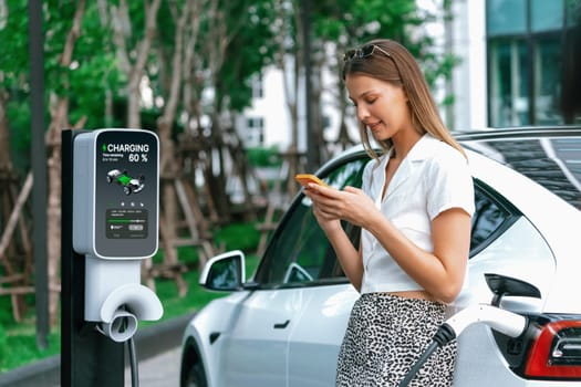 Young woman travel with EV electric car charging in green sustainable city outdoor garden in summer. Urban sustainability lifestyle by green clean rechargeable energy of electric BEV vehicle innards