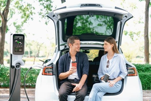 Lovely young couple drinking coffee while recharging battery for electric car during road trip travel EV car in natural forest or national park. Eco friendly travel during vacation and holiday. Exalt