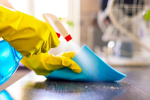 Close-up of hand in yellow gloves cleaning a wooden surface with a spray bottle and blue cloth, hygiene in the household.