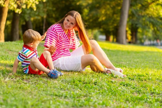 A cheerful young mother engages in play with her toddler son on a sunlit grassy park during summer.