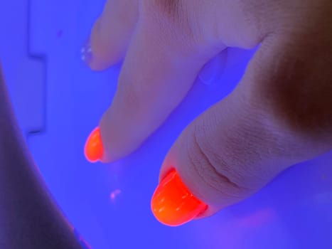 Female hand with neon orange manicure inside a blue UV lamp, drying gel polish. Manicure process, drying stage. High quality photo