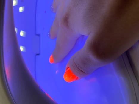 Female hand with neon orange manicure inside a blue UV lamp, drying gel polish. Manicure process, drying stage. High quality photo