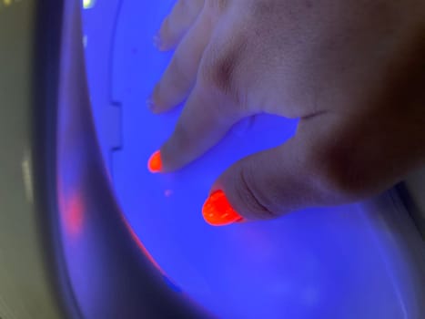 Female hand with neon orange manicure inside a blue UV lamp, drying gel polish. Manicure process, drying stage. High quality photo