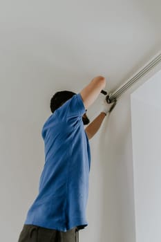One young Caucasian guy, brunette in a blue T-shirt, stands half-sided on a chair and unscrews a bolt from the cornice with a screwdriver to dismantle it, close-up view from below.