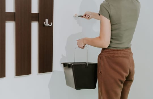 One young unrecognizable Caucasian brunette woman stands sideways, holds a bucket of paint and paints a wall with white paint with a brush near the wardrobe, close-up side view.