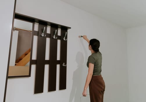 One young beautiful Caucasian brunette woman stands sideways near a wardrobe for outerwear with a mirror and paints the wall with white paint with a brush, close-up side view.