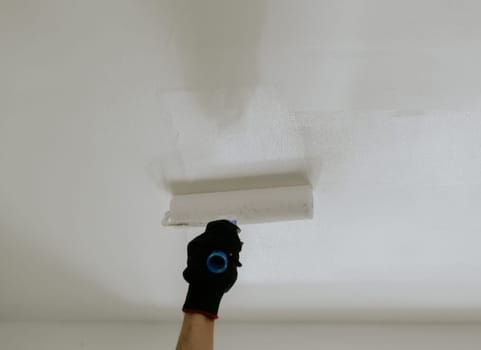 The hand of a young unrecognizable man paints the ceiling with white paint using a roller, bottom side view close-up with selective focus