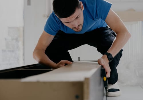 One young handsome Caucasian man in a blue T-shirt and black pants is repairing a broken back wall of an old wardrobe during the day in the kitchen, close-up side view.