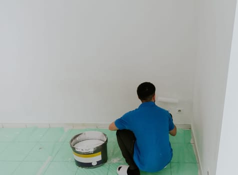 One young handsome dark-haired Caucasian man in a blue T-shirt is squatting on a tiled floor with a green oilcloth from his back holding and using a roller to paint a wall with white paint, side view close-up with selective focus.