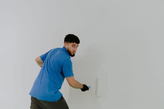 One young handsome dark-haired bearded Caucasian man in a blue T-shirt stands with his back tilted, looking at the camera and painting a wall with a roller of white paint, side view close-up with selective focus.