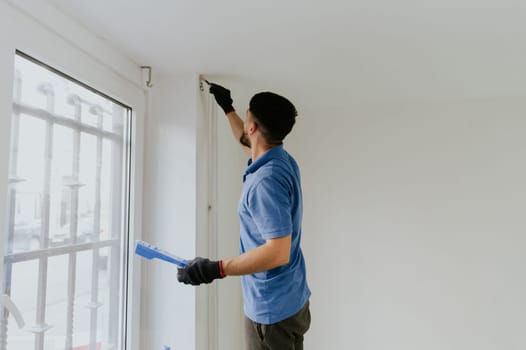 One young handsome dark-haired bearded Caucasian man in a blue T-shirt stands sideways by the window and paints the corners of the wall under the ceiling with a brush with white paint, close-up side view with selective focus.