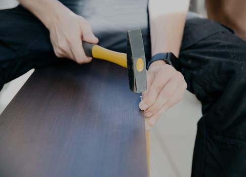 One young unrecognizable Caucasian man in a blue T-shirt and dirty black pants hammers a nail with a yellow hammer into the broken back wall of an old brown cabinet, close-up side view.