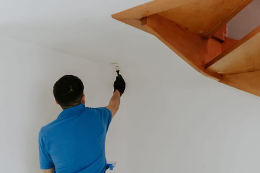 One young handsome dark-haired Caucasian man in a blue T-shirt stands from the back and paints with a brush with white paint the corners of the wall under the wooden stairs, close-up side view with selective focus.