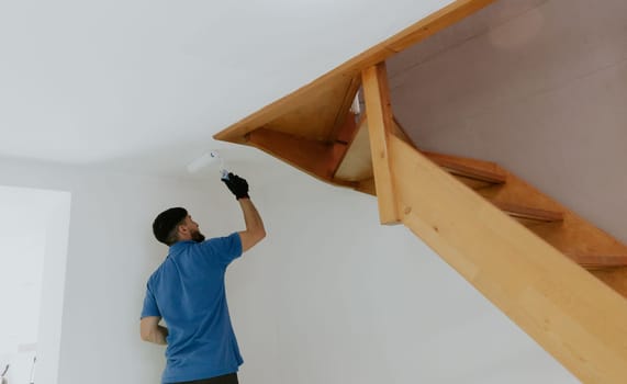 One young handsome dark-haired Caucasian man in a blue T-shirt stands from behind and paints with a roller the ceiling under a wooden staircase next to a doorway, bottom side close-up view with selective focus.