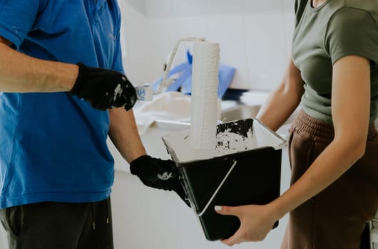 One young Caucasian unrecognizable couple is doing renovations: the girl is holding a bucket of paint, and the guy is dipping a roller into it, standing in the kitchen on a summer day, close-up view with selective focus.