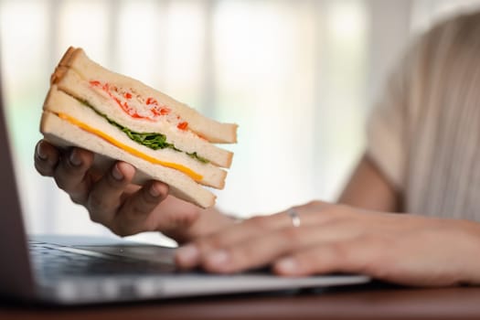 Close-up of a woman using a laptop while holding a tasty sandwich for the concept of fast food eating at the workplace