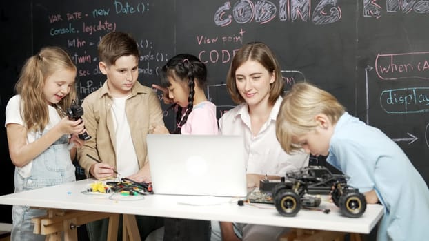 Caucasian teacher using laptop and explain about engineering system while diverse student holding electronic equipment in front of blackboard with prompt or code written in STEM class. Erudition.