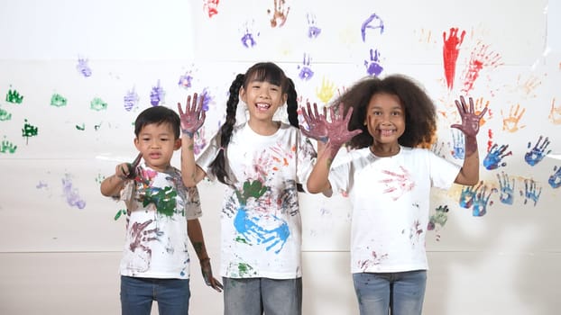 Diverse students put hands up together show colorful stained hands. Group of multicultural learner standing at white background with stained hands while looking at camera in lively mood. Erudition.