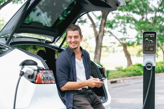 Man using smartphone online banking application to pay for electric car battery charging from EV charging station during vacation holiday road trip at national park or summer forest. Exalt