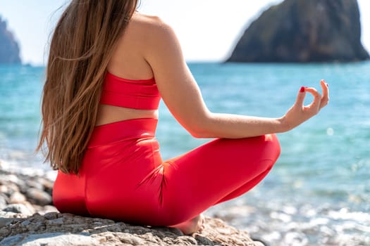 A young woman in red leggings and a red top with long loose hair practices yoga outdoors by the sea on a sunny day. Women's yoga, fitness, Pilates. The concept of a healthy lifestyle, harmony