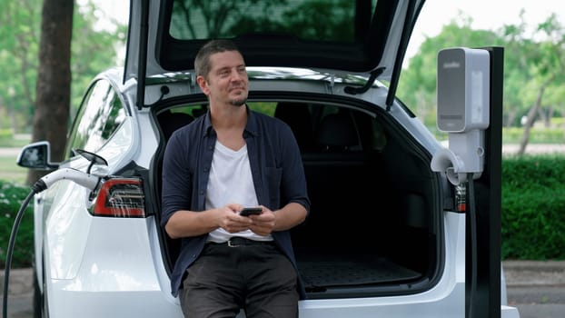 Man using smartphone online banking application to pay for electric car battery charging from EV charging station during vacation holiday road trip at national park or summer forest. Exalt