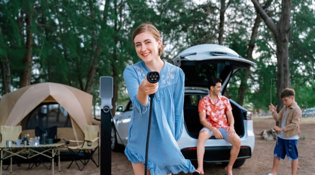 Outdoor adventure and family vacation camping at sea travel by eco friendly car. Cheerful woman or mother holding, pointing EV charger point with playful and happiness posture in campsite. Perpetual