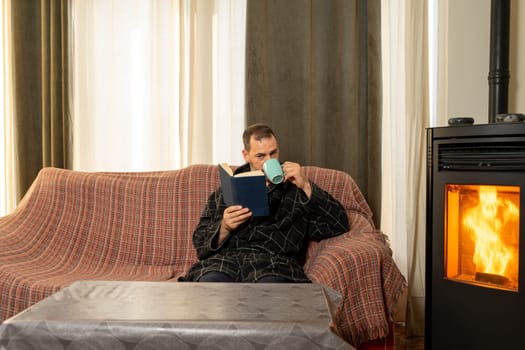 Hispanic man with a beard in his 40s wearing a bathrobe drinking coffee and reading relaxedly on the couch at home in the heat of a pallet stove