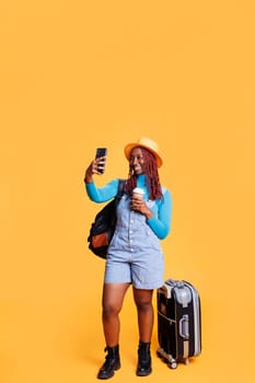 Traveller taking photos on vacation trip, making silly faces on smartphone and drinking coffee. Female person having fun with pictures in studio, carrying backpack and trolley bags.