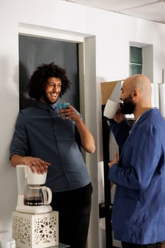 Two smiling arab businessmen having coffee break and talking in business office. Company employees standing with tea mugs while laughing and chatting in coworking space workplace