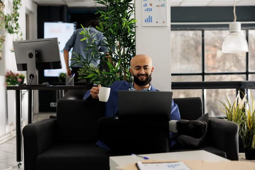 Smiling relaxed startup business employee drinking coffee and working on laptop in office. Happy arab man sitting on couch and holding tea mug while checking successful marketing results
