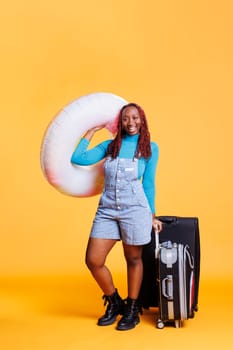 African american girl leaving on holiday with inflatable ring and trolley bags, feeling excited about vacation. Young woman enjoying weekend activities abroad, carrying luggage.