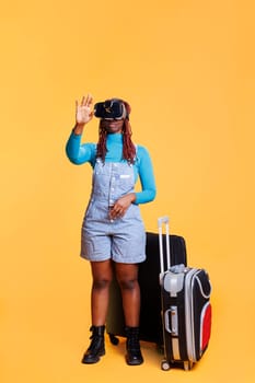 Young adult having fun with vr glasses in studio, posing over orange background with virtual reality headset and trolley bags. Female traveller using modern goggles with 3d vision.