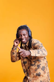 Joyful young adult enjoying radio song on headphones in studio over yellow background, dancing and having fun during free time. African american man relaxing and listening to audio on camera