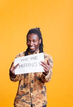 Company worker holding paper sign to make job offer in front of camera, hiring perfect candidate for career opportunity. African american employee asking people to attend job interview in studio