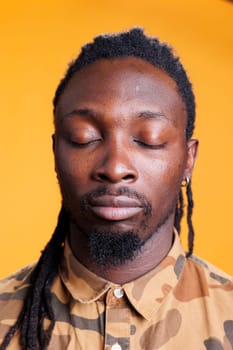 Relaxed man breathes deeply in studio, meditating with concentrated thoughts over yellow background. Concentrated african american person trying to relax, practicing wellness exercise.