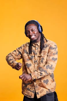 African american man listening music using headphones, dancing alone in studio over yellow background. Portrait of smiling young adult enjoying joyful playlist, having fun on song rhythm