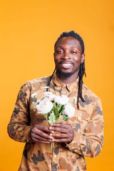 Smiling man posing with white roses bouquet, holding romantic gift for girlfriend during valentine s day. African american person celebrating love holiday in studio over yellow background