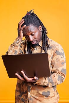 Pensive african american man brainstorming business ideas, checking marketing strategy using laptop computer in studio. Thoughtful young adult holding digital device, browsing on internet