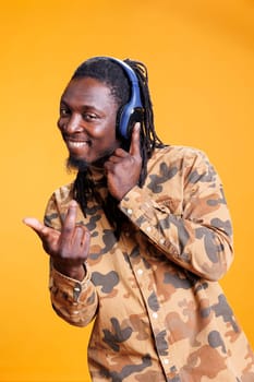 Smiling young adult enjoying radio music on headphones, standing over blue background. African american man listening to mp3 song, dancing and feeling happy using wireless headphones