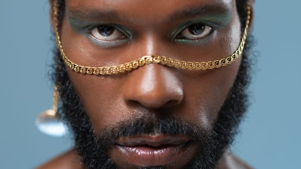 Closeup portrait of bearded African American gay man with blue eyeshadow and golden accessory on face. Serious homosexual adult male with makeup looking at camera on blue background.