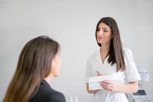Confident female cosmetologist writing while talking with woman client. Female professional beauty doctor in uniform listening to customer. Dermatologist and patient in clinic.