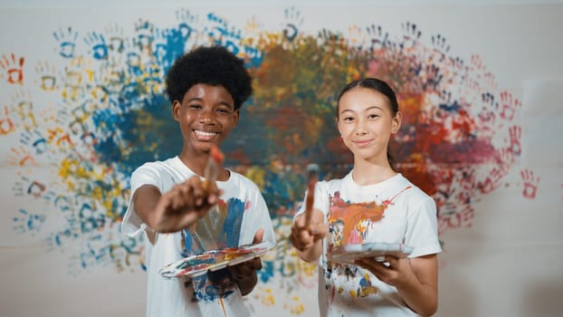 Highschool student with mixed races standing and looking at camera at art lesson. Diverse children hold the paintbrush while pose with confident at colorful stained wall. Creative style. Edification.